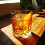 A glass of Kikori Rice Whiskey cocktail with ice and an orange slice, bathed in sunlight.