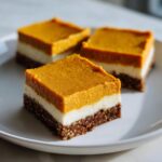 Three square slices of layered Pumpkin Cheesecake Bars featuring a dark crust, white filling, and bright orange pumpkin topping.