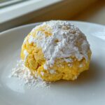 A single, bright yellow Lemon Cake Mix Cookies, heavily dusted with white powdered sugar, resting on a white plate.