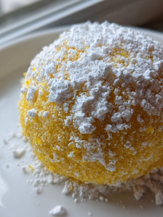 A single, bright yellow Lemon Cake Mix Cookie dusted heavily with white powdered sugar, resting on a white plate.