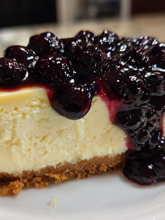 A rich slice of lemon cheesecake topped generously with dark, glossy blueberry compote.
