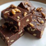 Close-up of rich, dark chocolate Maple Walnut Fudge squares topped with chopped walnuts on a white plate.