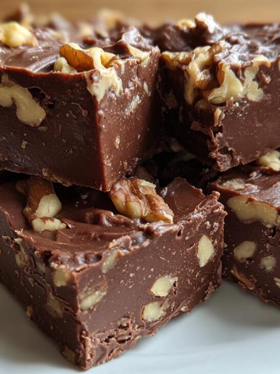 Close-up of rich, dark chocolate Maple Walnut Fudge squares stacked on a white plate, topped with visible walnut pieces.