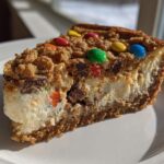 A close-up slice of Monster Cookie Cheesecake showing a creamy filling, cookie crust, and colorful M&M candies on top.