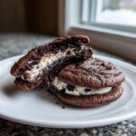 Two rich, dark chocolate Oreo Cheesecake Cookies on a white plate, one broken open to show the creamy white filling with cookie chunks.