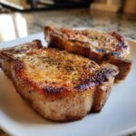 Two perfectly cooked oven baked bone in pork chops seasoned with herbs and spices, served on a white plate.