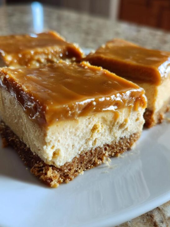 Three squares of rich Peanut Butter Cheesecake Squares topped with glossy caramel sauce on a white plate.