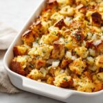 Close-up of a white baking dish filled with Perfect Homemade Stuffing, featuring golden-brown bread cubes, onions, and herbs.