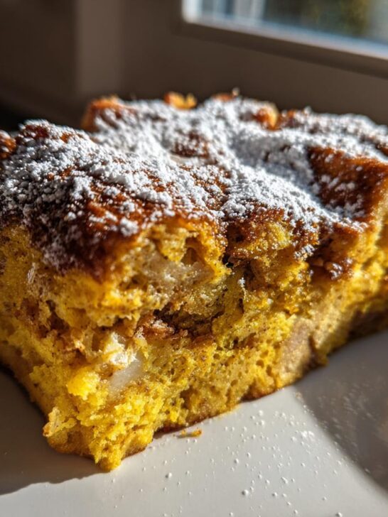 A close-up of a golden slice of Pumpkin French Toast Casserole topped with powdered sugar, sitting on a white plate.