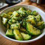 A close-up of a Refreshing Cucumber Avocado Salad in a white bowl, topped with fresh herbs and seasoning.