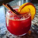 A festive red cranberry cocktail garnished with an orange slice and cinnamon sticks, perfect for Cold Weather Weekend Cocktails.