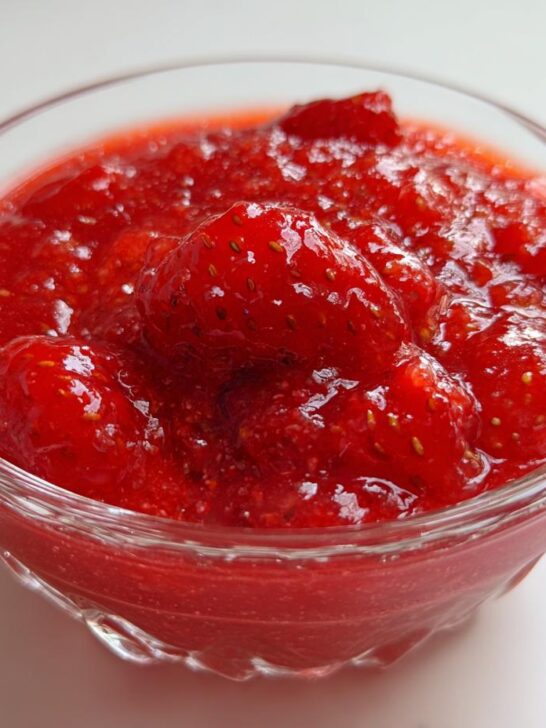 A close-up of a clear glass bowl filled with vibrant, chunky strawberry sauce, showcasing whole and halved strawberries.