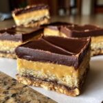 A close-up, tasty shot of a square Peanut Butter Nanaimo Bars showing the three distinct layers: crumbly base, thick peanut butter filling, and chocolate topping.
