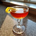 A close-up of The Paper Plane cocktail in a coupe glass, garnished with a lemon twist.