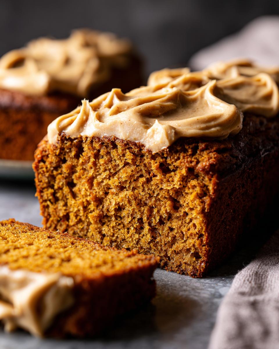 A slice of moist vegan pumpkin bread topped with creamy maple chai frosting, with another loaf in the background.