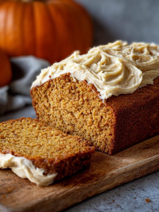 A slice of moist Vegan Pumpkin Bread topped with creamy Maple Chai Frosting, with a whole loaf and pumpkins in the background.