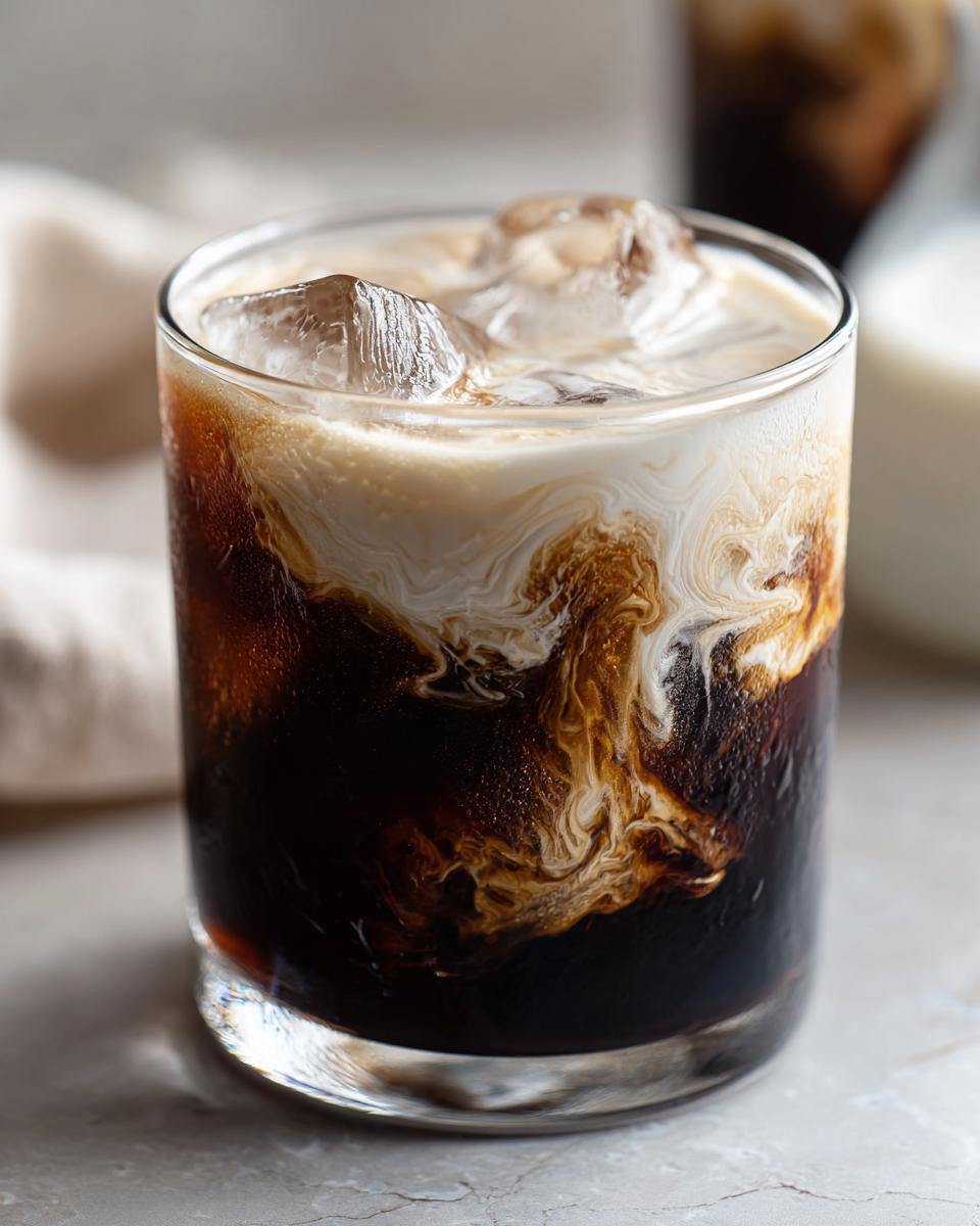 Close-up of a perfectly made White Russian cocktail with swirling cream, dark coffee liqueur, and ice cubes.