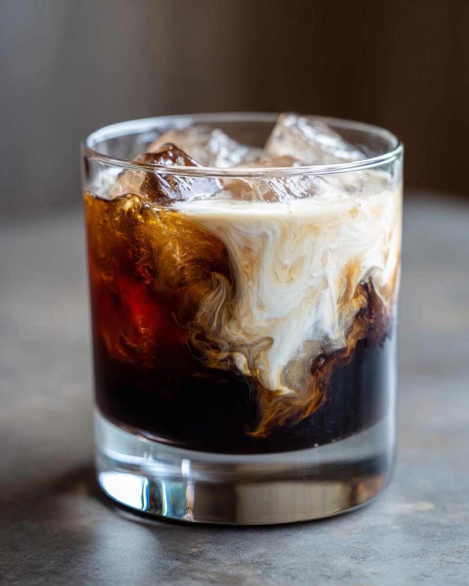 A close-up of a White Russian cocktail in a glass, showing the distinct layers of coffee liqueur and cream swirling together over ice.