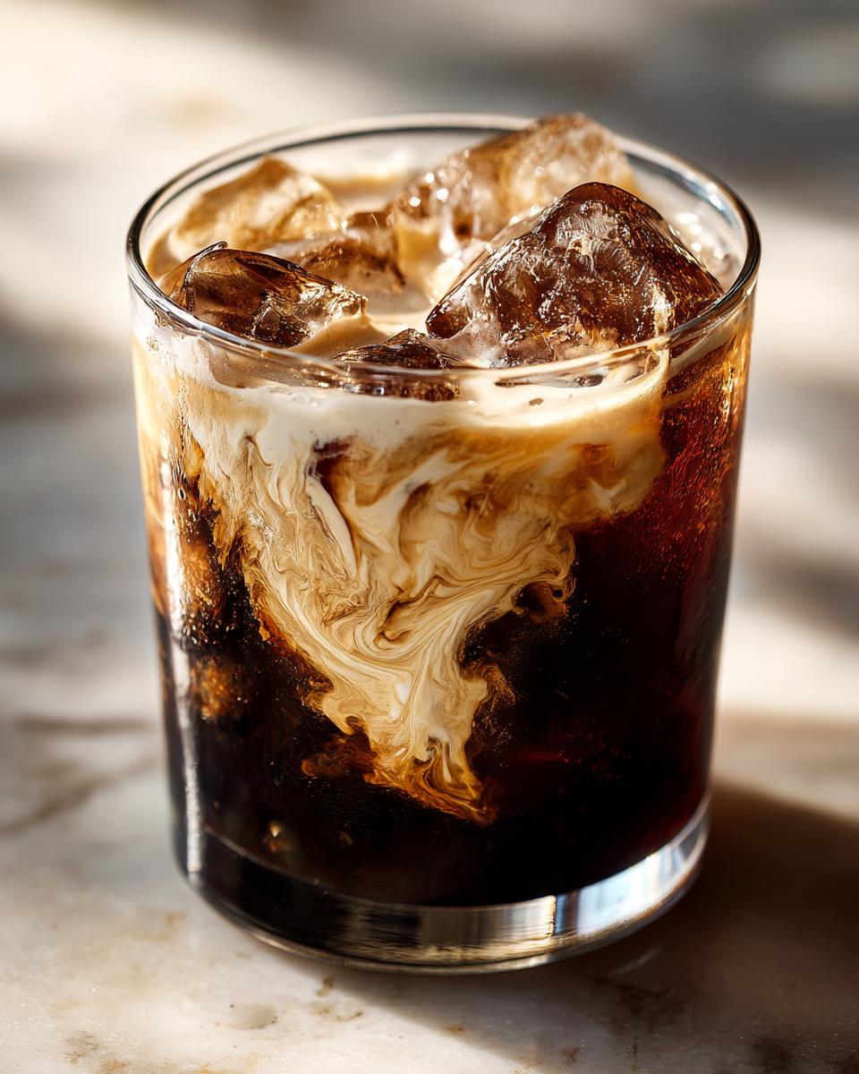 A close-up shot of a White Russian cocktail with swirling cream and ice cubes in a glass.