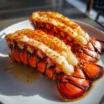 Two cooked lobster tails, split open to show white meat seasoned with paprika, resting on a white plate. This is the result of Air Fryer Lobster.