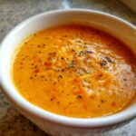 Close-up of creamy orange Apple Cheddar Sweet Potato Soup topped with herbs and melted cheese.