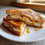 Two halves of golden brown Banana Bread Stuffed French Toast filled with cream cheese and banana slices, drizzled with syrup.