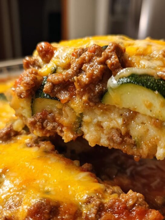 A close-up of a serving being lifted from a Beef Zucchini Casserole, showing layers of ground beef, zucchini slices, and melted cheddar cheese.