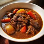 Close-up of rich, dark stew featuring chunks resembling meat, carrots, and potatoes in a savory broth: The Best Plant Based Jamaican Oxtail.