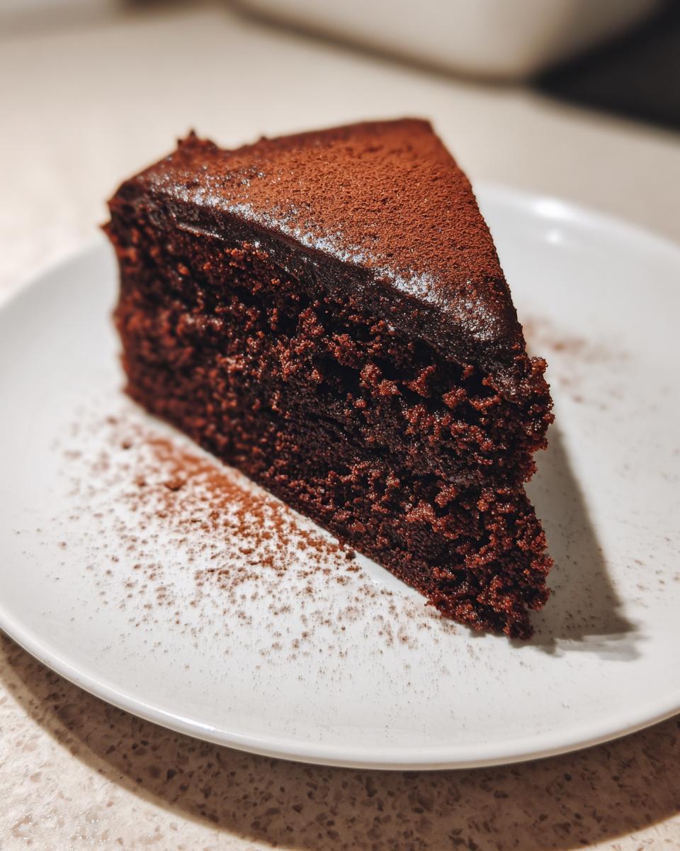 Close-up of a rich, dark slice of The Best Vegan Chocolate Cake dusted with cocoa powder on a white plate.