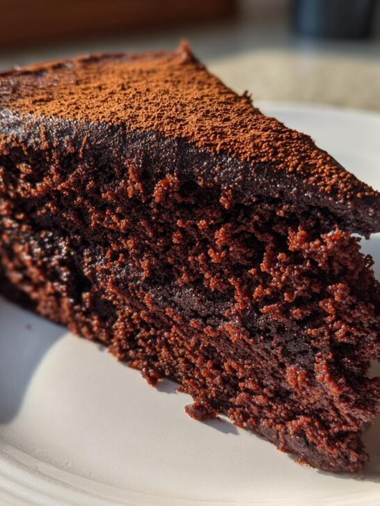 A close-up slice of The Best Vegan Chocolate Cake, showing moist crumb and cocoa powder topping on a white plate.