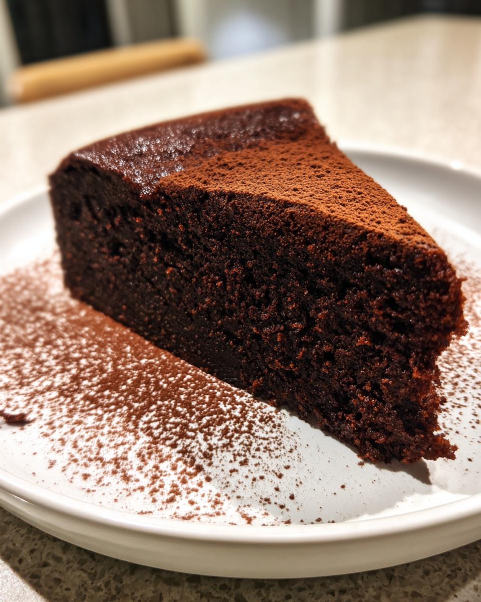 A rich, dark slice of The Best Vegan Chocolate Cake dusted generously with cocoa powder on a white plate.