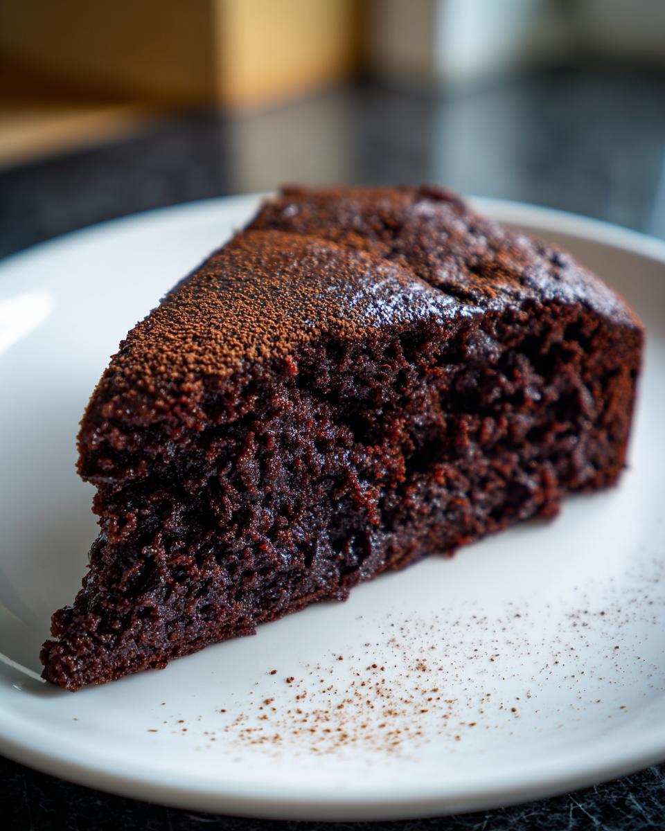 Close-up of a rich, dark slice of The Best Vegan Chocolate Cake dusted with cocoa powder on a white plate.