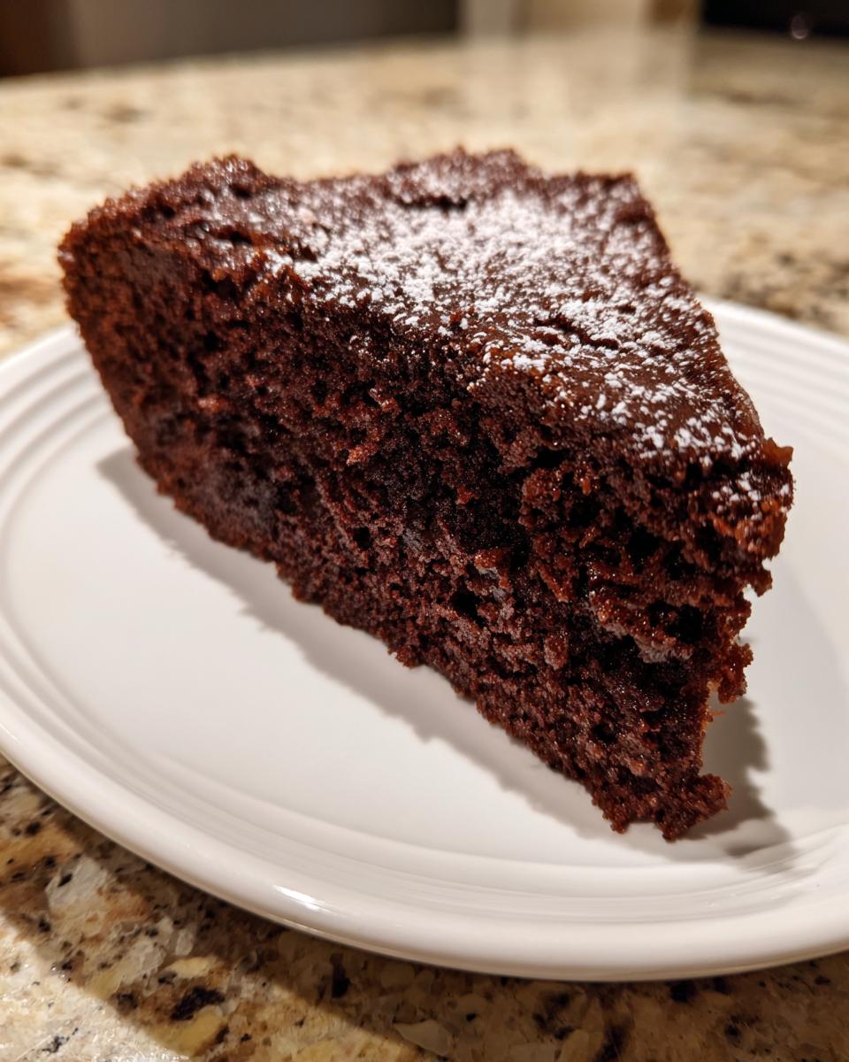 A moist slice of The Best Vegan Chocolate Cake dusted lightly with powdered sugar, served on a white plate.