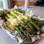 Close-up of grilled asparagus spears topped with melted cheese, served in aluminum foil for Cheesy Grilled Asparagus Foil Packs.