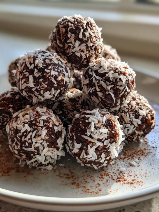 A stack of homemade Chocolate Coconut Energy Bites coated in shredded coconut, sitting on a plate dusted with cocoa powder.