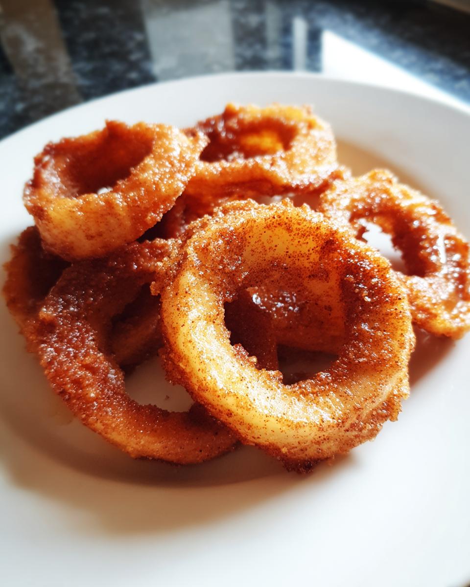 A pile of golden brown, sweet Apple Rings dusted with cinnamon sugar served on a white plate.