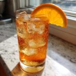 A tall glass filled with a bubbly, amber Citrus Honey Mocktail over ice, garnished with a bright orange slice.