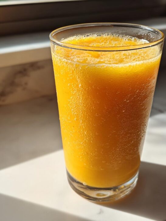 A tall glass filled with a bright orange Coconut Mango Mocktail, showing condensation on the outside, sitting on a sunny counter.