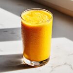 A tall glass filled with a vibrant, frothy, orange Coconut Mango Mocktail, showing condensation beads on the outside.