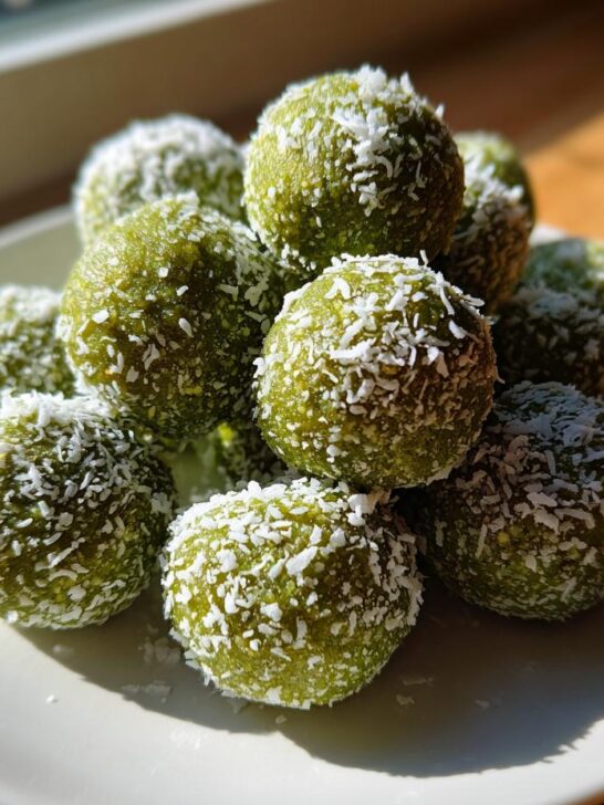 A close-up of a pile of vibrant green Coconut Matcha Energy Bites rolled in shredded coconut, sitting on a white plate.