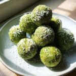 A stack of bright green Coconut Matcha Energy Bites rolled in shredded coconut on a light plate.