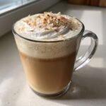 A clear glass mug filled with a layered Coconut Vanilla Latte, topped with whipped cream and toasted coconut flakes.