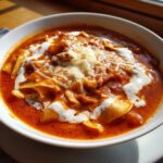 Close-up of a bowl of rich, red Comforting Vegetarian Lasagna Soup topped with dollops of white cream and shredded melted cheese.