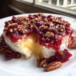 Close-up of baked brie oozing warm cheese, topped with vibrant cranberry sauce and crunchy pecans.