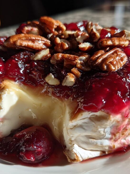 Close-up of warm, gooey baked brie topped with bright red cranberry sauce and crunchy pecans, a slice removed.