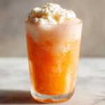 Close-up of a tall glass filled with a bright orange, fizzy Creamsicle Ice Cream Sodas, topped generously with whipped cream.