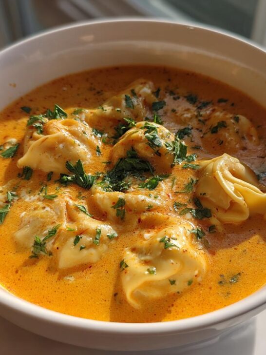 A close-up of a white bowl filled with rich, orange Creamy Cajun Pumpkin Tortellini Soup topped with fresh parsley.