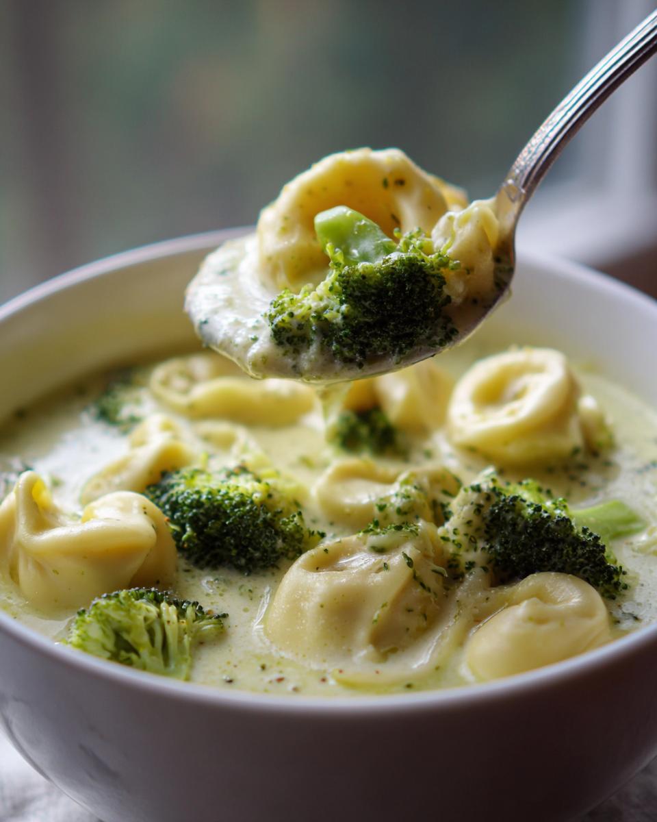 A spoonful of creamy lemon broccoli tortellini soup lifted from a white bowl, showing cheese tortellini and bright green broccoli florets.