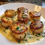 Six golden-brown seared scallops swimming in a rich, creamy lemon butter sauce garnished with fresh parsley.