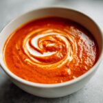 Close-up of a white bowl filled with rich, orange-red Creamy Roasted Garlic Tomato Soup, swirled with cream.